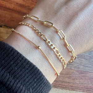 May include: Three gold bracelets of different styles are worn on a wrist. The bracelets include a box chain, a Figaro chain, and a paperclip chain. The bracelets are displayed on a person's wrist, with a black sleeve in the background.