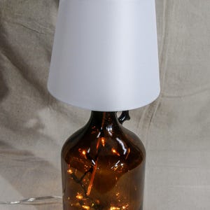 Könnte beinhalten: Dekorative Tischlampe mit einem braunen Glasflaschenfuß und einem weißen Lampenschirm. Die Flasche ist mit kleinen, warmweißen LED-Lichtern gefüllt. Die Lampe ist für den Innenbereich konzipiert.