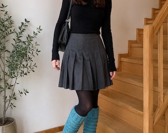 May include: A woman wearing a black long-sleeved sweater and a gray pleated skirt. She is also wearing black tights, black patent leather shoes, and turquoise leg warmers. A black purse hangs from her shoulder. The background includes a wooden staircase and a potted plant.