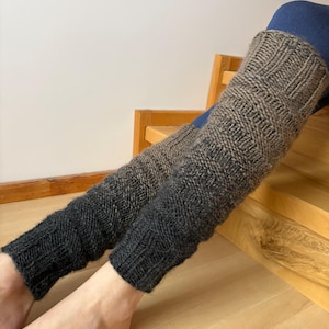 May include: A pair of knitted leg warmers in a gradient of gray and black. The leg warmers are made of a textured yarn and are worn on the legs. The top portion is a lighter gray, transitioning to a darker gray and then black at the bottom.