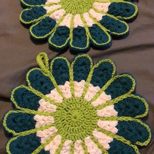 Pode incluir: Dois pegadores de panela de crochê em forma de flor. Os pegadores são verdes, brancos e rosa.