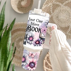 May include: A white tumbler with a handle, featuring the text "Just One More Book" in black script. The design includes watercolor-style flowers, butterflies, and book illustrations in shades of purple and gray. A straw is visible.