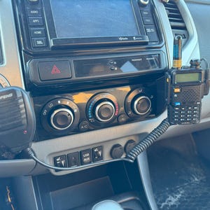 May include: Close-up of a car's dashboard with a black radio, climate control knobs, and a black two-way radio with a silver antenna mounted on the right side.