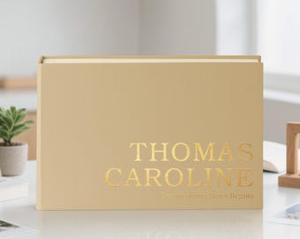 May include: A beige wedding album with gold lettering that reads "THOMAS CAROLINE" and "Our Wedding Story Begins." The album is rectangular and sits on a white surface. A small potted plant is visible in the background.