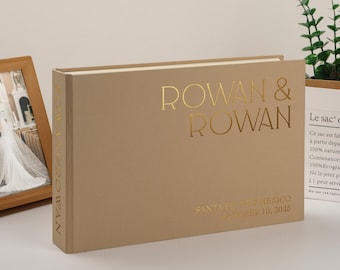 May include: A beige wedding guest book with gold lettering that reads "ROWAN & ROWAN". The book also has the text "SANT VER ME MEXICO OCTOBER 10, 2025". A framed photo and greenery are in the background.