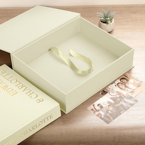 May include: A cream-coloured gift box with an open lid and a satin ribbon inside. A book with gold lettering reading "Edward & Charlotte" is next to the box. Two photographs and a small succulent plant are also visible.