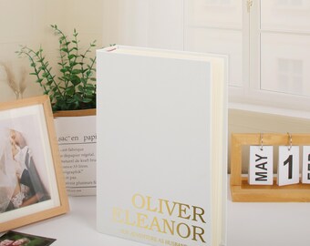 May include: A white photo album with gold lettering that reads "OLIVER ELEANOR OUR ADVENTURE AS HUSBAND & WIFE." The album is standing upright on a white surface. A framed photo and a wooden calendar are in the background.