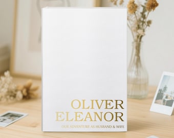 May include: A white photo album with gold lettering that reads "OLIVER ELEANOR" and "OUR ADVENTURE AS HUSBAND & WIFE." The album is on a wooden surface, with a vase of dried flowers and framed art in the background.