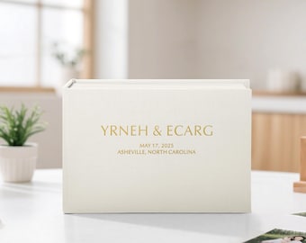 May include: A cream-colored photo album with gold lettering. The text on the front reads "YRNEH & ECARG", "MAY 17, 2025", and "ASHEVILLE, NORTH CAROLINA". The album is rectangular and sits on a white surface.