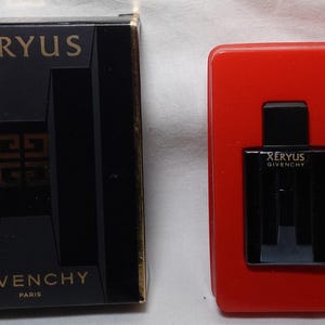 Puede incluir: Una caja negra con letras doradas que dicen "Xeryus Givenchy Paris". La caja contiene un pequeño frasco de perfume con la misma marca. El frasco se muestra sobre un fondo rojo con un pequeño alfiler de velero.