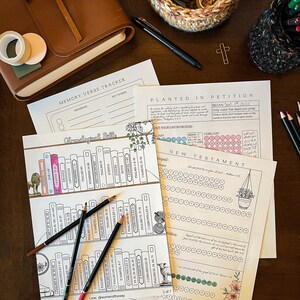 May include: A collection of stationery items on a dark wooden surface. Includes printed Bible study pages, colored pencils, a leather journal, washi tape, and a pen. The pages feature text and illustrations, with the text "Memory Verse Tracker" and "Chronological Bible".