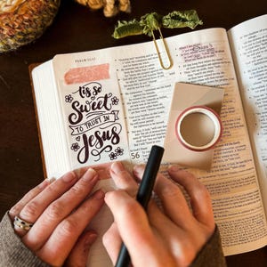 Può includere: Una Bibbia aperta con un segnalibro decorativo che recita "'Tis so Sweet to Trust in Jesus". Sono visibili anche un rotolo di nastro decorativo e una penna. L'immagine è ripresa dall'alto, con una calda e invitante tavolozza di colori.