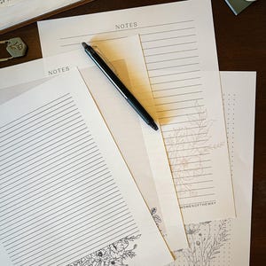 Puede incluir: Varias hojas de papel con la palabra "NOTES" impresa, algunas rayadas y otras con dise&ntilde;os florales. Un bol&iacute;grafo negro descansa sobre las p&aacute;ginas. Una hoja presenta un vers&iacute;culo b&iacute;blico en la parte inferior. Los papeles est&aacute;n sobre una superficie de madera oscura.