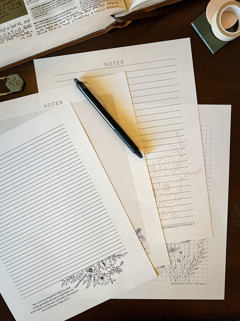 Puede incluir: Varias hojas de papel con la palabra "NOTES" impresa, algunas rayadas y otras con dise&ntilde;os florales. Un bol&iacute;grafo negro descansa sobre las p&aacute;ginas. Una hoja presenta un vers&iacute;culo b&iacute;blico en la parte inferior. Los papeles est&aacute;n sobre una superficie de madera oscura.