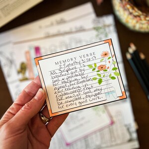 May include: A handwritten memory verse card with the text "2 Timothy 3:16-17" and floral illustrations. The card is held in a hand, with other cards and colored pencils in the background. The card has an orange border.