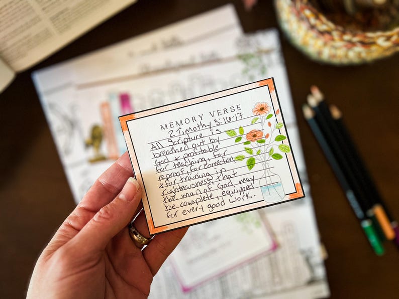 May include: A handwritten memory verse card with the text "2 Timothy 3:16-17" and floral illustrations. The card is held in a hand, with other cards and colored pencils in the background. The card has an orange border.