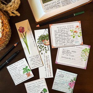 Puede incluir: Una colecci&oacute;n de marcap&aacute;ginas y tarjetas hechas a mano con ilustraciones florales y bot&aacute;nicas, junto con texto escrito a mano. Los art&iacute;culos est&aacute;n dispuestos sobre una superficie de madera oscura, con un cuaderno y bol&iacute;grafos. El tema es de fe e inspiraci&oacute;n.