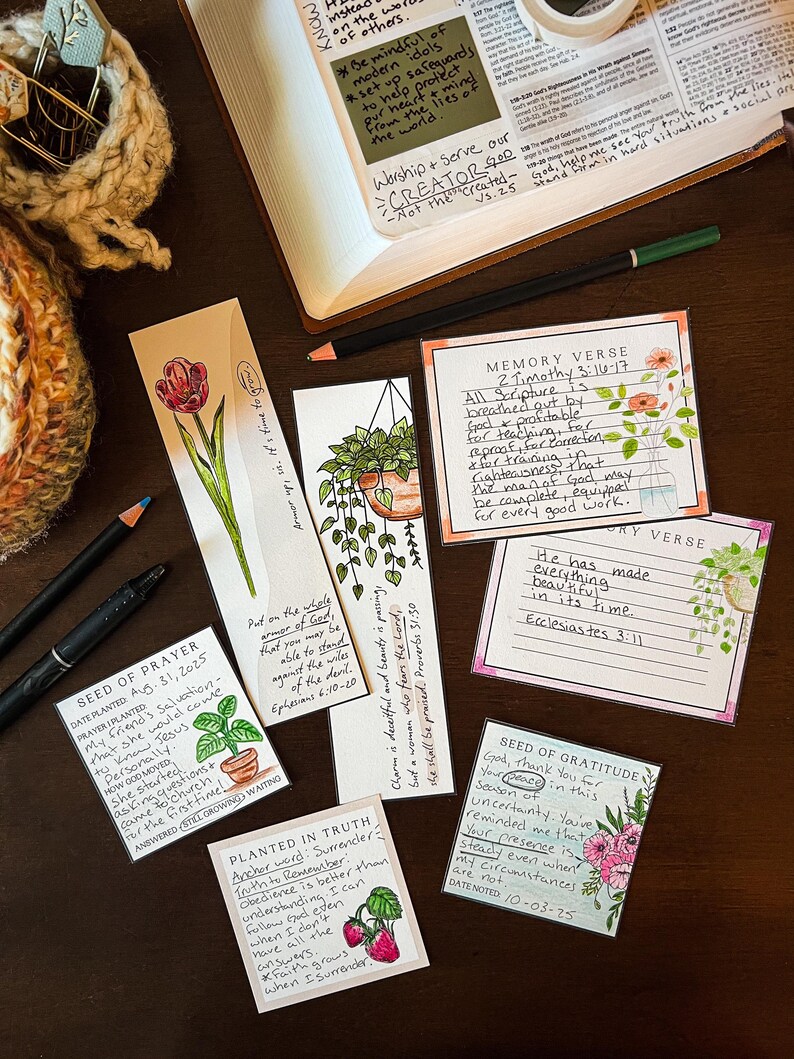 Puede incluir: Una colecci&oacute;n de marcap&aacute;ginas y tarjetas hechas a mano con ilustraciones florales y bot&aacute;nicas, junto con texto escrito a mano. Los art&iacute;culos est&aacute;n dispuestos sobre una superficie de madera oscura, con un cuaderno y bol&iacute;grafos. El tema es de fe e inspiraci&oacute;n.