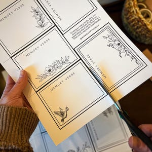 May include: A person is cutting out memory verse cards with scissors. The cards are printed on white paper with black floral illustrations and the text "MEMORY VERSE". The instructions and the @WOMENOFTHEWAY are also printed on the paper.