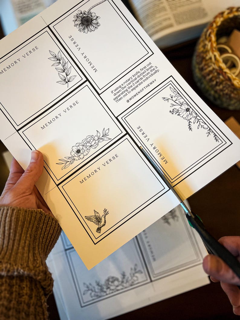 May include: A person is cutting out memory verse cards with scissors. The cards are printed on white paper with black floral illustrations and the text "MEMORY VERSE". The instructions and the @WOMENOFTHEWAY are also printed on the paper.