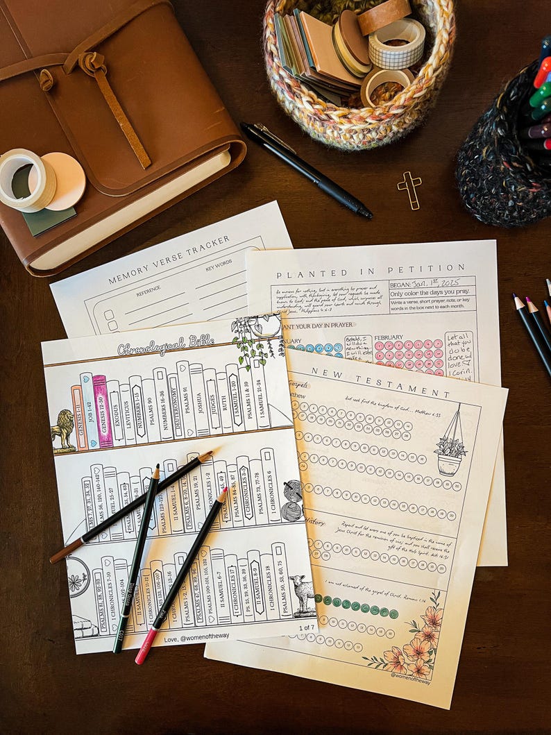 May include: A collection of stationery items on a dark wooden surface. Includes printed Bible study pages, colored pencils, a leather journal, washi tape, and a pen. The pages feature text and illustrations, with the text "Memory Verse Tracker" and "Chronological Bible".