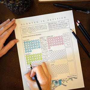 May include: A white paper titled "Planted in Petition" with a calendar layout for prayer journaling. The calendar is being colored with a black pencil. The paper includes text and floral illustrations. Colored pencils and a pen are also visible.