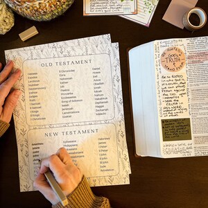 May include: A Bible study setup with a printed list of Old and New Testament books, a Bible, and decorative stationery. The image includes a handwritten memory verse and the words "ROOTED IN GOD'S TRUTH STAND FIRM."