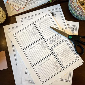 May include: A collection of white paper memory verse cards with black borders and floral illustrations. The cards are arranged on a dark wooden surface, alongside a pair of scissors, an open book, and a pen. Some cards have handwritten notes.