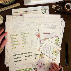 Op de afbeelding: Een verzameling gedrukte Bijbelstudiepagina's met bloemenillustraties, waaronder een geheugenverstracker en een petitieblad. Een Bijbel, pennen en washitape zijn ook zichtbaar, wat een studie-thema-opstelling creëert.