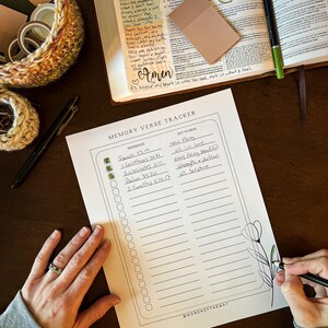 May include: A Memory Verse Tracker with a floral design is being filled out with a black pen. The tracker includes sections for references and keywords. A Bible, sticky notes, and decorative basket are also visible.