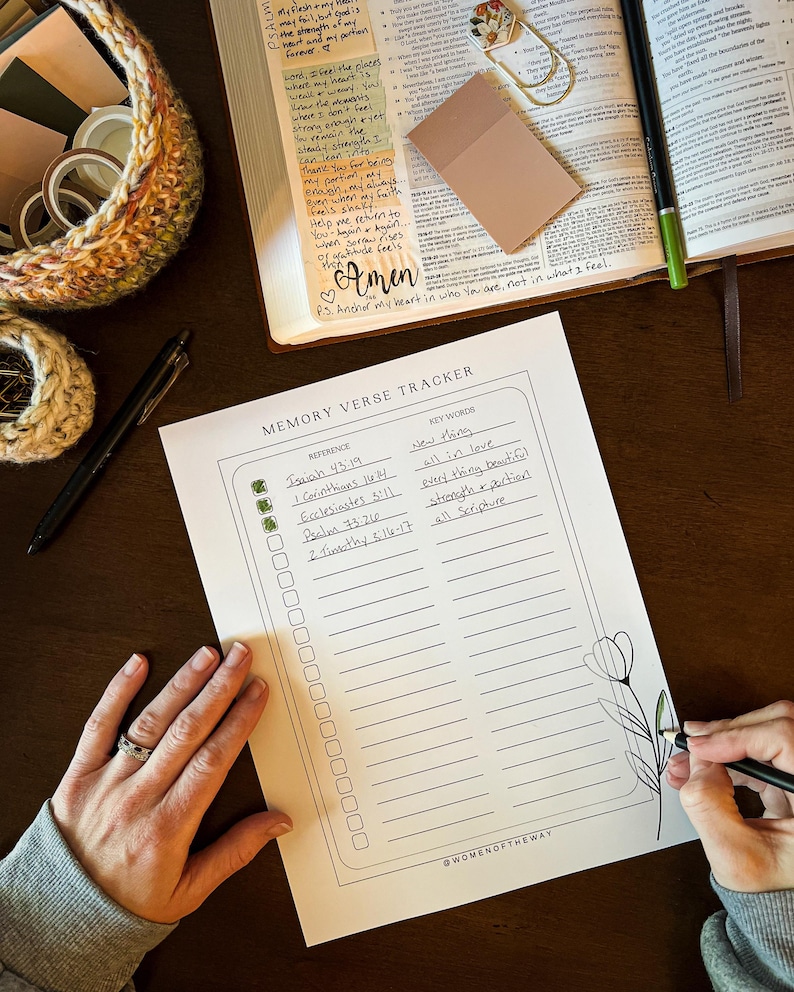 May include: A Memory Verse Tracker with a floral design is being filled out with a black pen. The tracker includes sections for references and keywords. A Bible, sticky notes, and decorative basket are also visible.