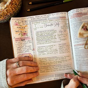 May include: An open Bible with handwritten notes, highlighted text, and colorful markers. The text includes the words "Hagar" and "Character Study." A hand with a ring is visible, holding the Bible. The word "EMBRACE" is written on a marker.