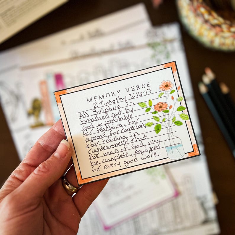 Puede incluir: Una mano sostiene una tarjeta de vers&iacute;culo de memoria con el texto "2 Timothy 3:16-17". La tarjeta tiene un borde naranja e ilustraciones florales. El texto dice "Toda la Escritura es inspirada por Dios..."