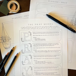 May include: A collection of printed prayer guides with the title "The Pray Model" and the text "Planted - Pray." The guides feature a layout for daily conversations with God, with sections for praise, repentance, asking, and yielding. Pencils, tape, and a pen are also visible.