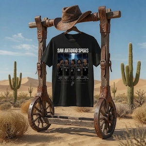 Pode incluir: Uma t-shirt preta de manga curta com uma impressão gráfica de cinco jogadores de basquetebol dos San Antonio Spurs e o texto "SAN ANTONIO SPURS" por cima. A t-shirt está num cabide de madeira suspenso numa estrutura de madeira rústica.