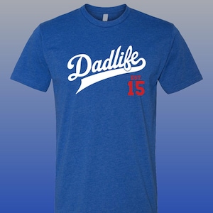 May include: Royal blue t-shirt featuring the white script text "Dadlife". Beneath this is "EST" and the number "15" in red. The shirt has a crew neck and short sleeves.