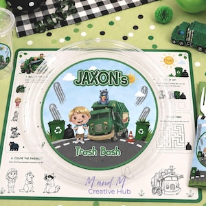 Könnte beinhalten: Ein Set aus transparentem Plastikbecher, Teller und Tischset mit Müllwagen-Motiv. Der Teller und das Tischset zeigen den Text "Jaxon's Trash Bash" mit Illustrationen eines Müllwagens, Zeichentrickfiguren und Mülleimern. Eine passende Serviette und ein Spielzeug-LKW sind ebenfalls vorhanden.