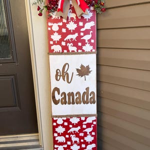 May include: A wooden sign with a red and white Canadian themed design. The sign says "Oh Canada" with a maple leaf. The sign is decorated with a burlap bow and red and white ribbon.