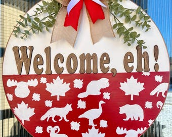 Könnte beinhalten: Runder Holz-Willkommensschild mit dem Text "Welcome, eh!" in braunen Buchstaben. Die obere Hälfte ist weiß, die untere rot mit weißen Tier- und Ahornblatt-Ausschnitten. Eine Schleife aus Jute und Rot mit Grünakzenten verziert die Oberseite.