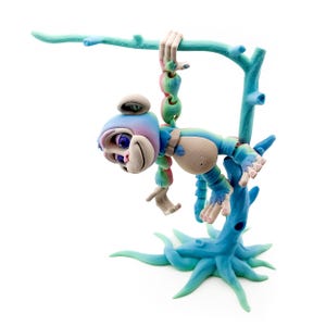 May include: A whimsical sculpture of a monkey-like creature in shades of blue, pink, and beige, hanging from a turquoise branch. The sculpture has a unique, cartoonish design with exaggerated features and is mounted on a turquoise base.