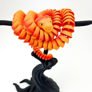 May include: A 3D-printed orange and red snake sculpture coiled around a black branch. The snake has a detailed head with yellow eyes and a textured body. The sculpture is set against a white background.