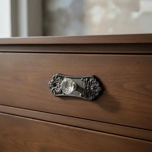 May include: Close-up of a dark brown wooden drawer with a decorative, antique-style handle. The handle features a clear crystal knob and a black, ornate backplate with floral detailing. The drawer is part of a larger wooden piece.