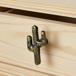 May include: A bronze-colored cactus-shaped drawer pull mounted on a light-colored wooden drawer. The drawer pull has a textured surface and is attached with a small, dark screw. The wood grain is visible on the drawer.