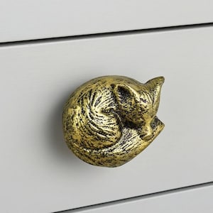 May include: A golden-colored cabinet knob in the shape of a curled-up fox. The knob has a textured surface and is mounted on a light gray cabinet door. The fox is in a sleeping position.