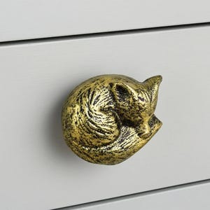 May include: A gold-colored, fox-shaped drawer pull. The fox is curled up in a sleeping position, with its tail wrapped around its body. The drawer pull is made of metal and has a textured surface.
