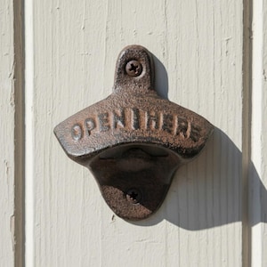 May include: A rustic, cast iron bottle opener mounted on a cream-colored wooden surface. The opener is a dark, weathered brown with the words "OPEN HERE" embossed across the top. Two screws secure it to the wood, and a shadow is cast to the right.