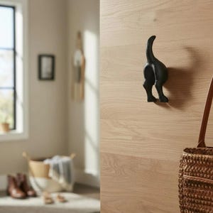 May include: A black cast iron wall hook shaped like a dog's rear end. The hook is mounted on a light wood-grain wall. A woven bag hangs nearby.