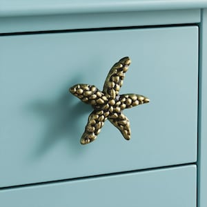 May include: A bronze-colored starfish-shaped drawer pull is mounted on a light blue cabinet. The drawer pull has a textured surface and is a decorative accent. The cabinet has a simple design.