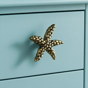 May include: A bronze-colored starfish-shaped drawer pull is mounted on a light blue cabinet. The drawer pull has a textured surface and is a decorative accent. The cabinet has a smooth, painted finish.