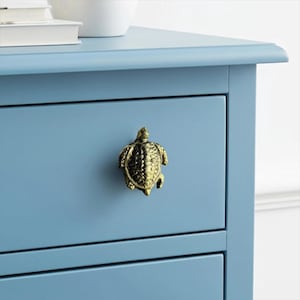 May include: A close-up of a light blue dresser drawer with a decorative brass turtle-shaped knob. The knob is centered on the drawer, and the dresser has a simple, classic design. The overall aesthetic is coastal or nautical.